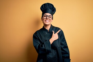 Young beautiful brunette chef woman wearing cooker uniform and hat over yellow background cheerful with a smile of face pointing with hand and finger up to the side with happy and natural expression