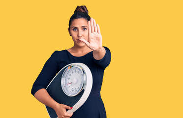 Young beautiful woman holding weighing machine with open hand doing stop sign with serious and confident expression, defense gesture