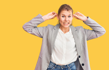 Young beautiful blonde woman wearing elegant jacket smiling pulling ears with fingers, funny...