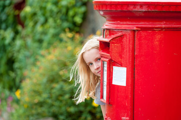 A young blonde girl peeks around a traditional red British Post Box