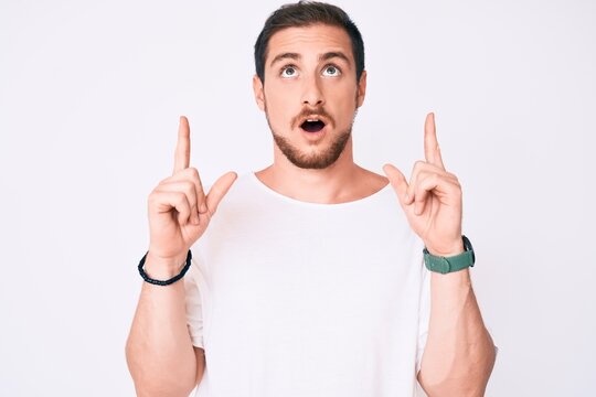 Young handsome man wearing casual white tshirt amazed and surprised looking up and pointing with fingers and raised arms.