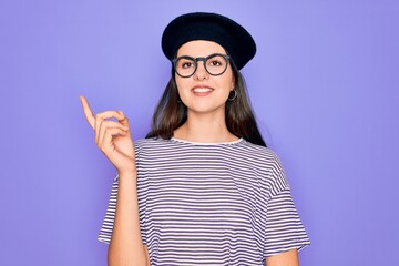 Young beautiful brunette woman wearing glasses and french beret over purple background with a big smile on face, pointing with hand and finger to the side looking at the camera.