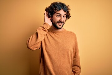 Young handsome man with beard wearing casual sweater standing over yellow background smiling with hand over ear listening an hearing to rumor or gossip. Deafness concept.