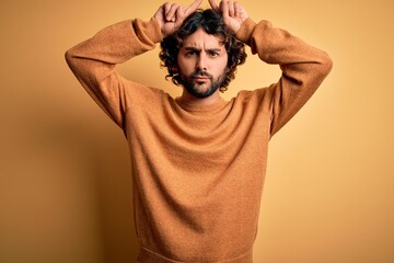 Young handsome man with beard wearing casual sweater standing over yellow background doing funny gesture with finger over head as bull horns