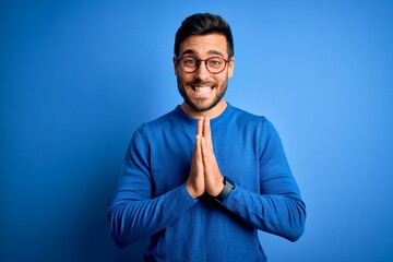 Young handsome man with beard wearing casual sweater and glasses over blue background praying with hands together asking for forgiveness smiling confident.