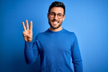 Young handsome man with beard wearing casual sweater and glasses over blue background showing and...