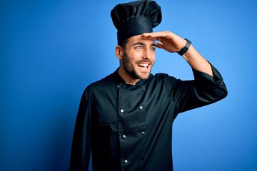 Young handsome chef man with beard wearing cooker uniform and hat over blue background very happy and smiling looking far away with hand over head. Searching concept.
