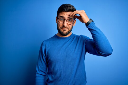 Young handsome man with beard wearing casual sweater and glasses over blue background worried and stressed about a problem with hand on forehead, nervous and anxious for crisis