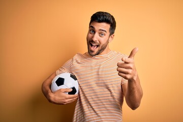 Handsome player man with beard playing soccer holding footballl ball over yellow background...