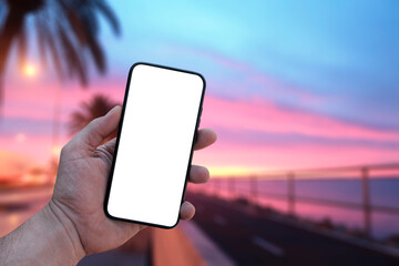 Mockup of the phone in the man hand with a white screen in his hand, against the background of the dawn of the sea and palm trees. Photo for travel and vacation on a tropical beach concept.