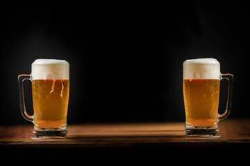 two cold mugs with beer, with overflowing foam, on wooden table and dark background, space for writing