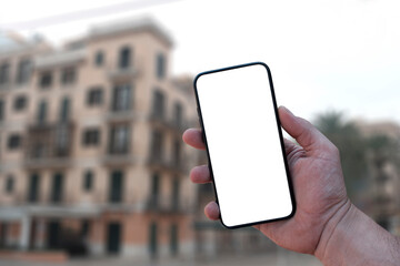 Mockup of the smartphone in the man hand, with a white screen on the background of urban buildings. Mobile shooting while traveling.