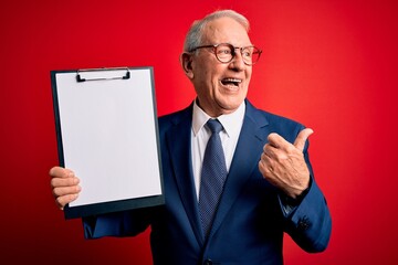 Senior grey haired business man holding clipboard over red background pointing and showing with thumb up to the side with happy face smiling