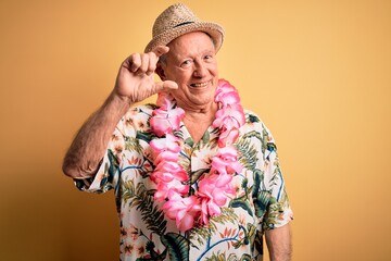 Grey haired senior man wearing summer hat and hawaiian lei over yellow background smiling and confident gesturing with hand doing small size sign with fingers looking and the camera. Measure concept.