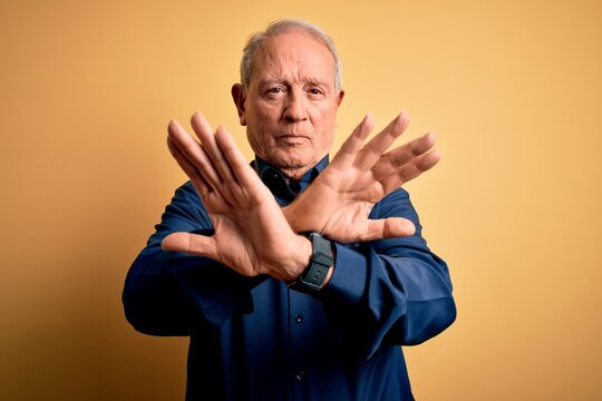 Grey haired senior man wearing casual blue shirt standing over yellow background Rejection expression crossing arms and palms doing negative sign, angry face