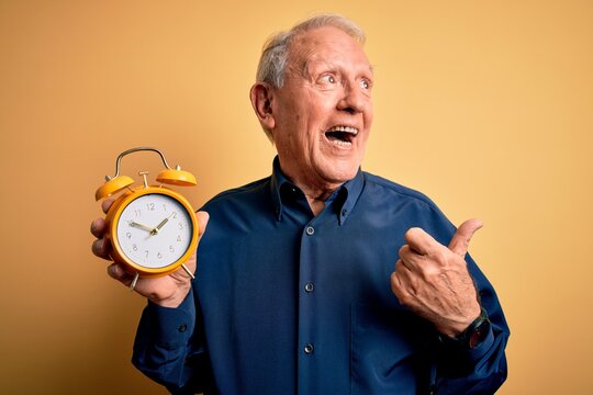 Senior grey haired man holding vintage alarm clock over yellow background pointing and showing with thumb up to the side with happy face smiling