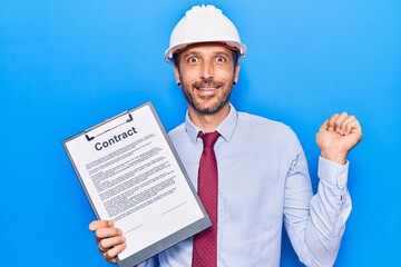 Young handsome man wearing architect hardhat holding contract document screaming proud, celebrating victory and success very excited with raised arm