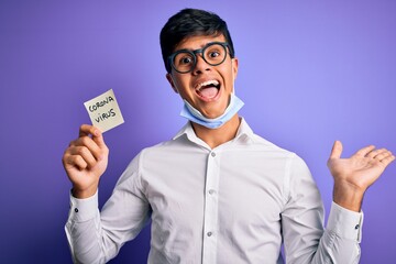 Young handsome man wearing medical mask holding reminder with corona virus message very happy and excited, winner expression celebrating victory screaming with big smile and raised hands