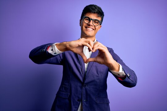 Young handsome business man wearing jacket and glasses over isolated purple background smiling in love doing heart symbol shape with hands. Romantic concept.