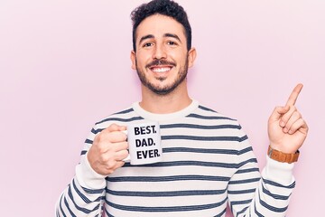Young hispanic man drinking mug of coffe with best dad ever message smiling happy pointing with hand and finger to the side