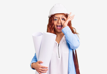Young latin woman wearing architect hardhat and leather bag holding blueprints smiling happy doing ok sign with hand on eye looking through fingers