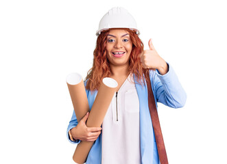 Young latin woman wearing architect hardhat holding blueprints smiling happy and positive, thumb up doing excellent and approval sign