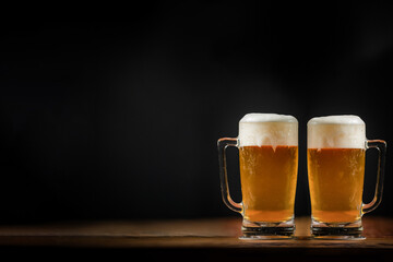 two cold mugs with beer, with overflowing foam, on wooden table and dark background, space for writing