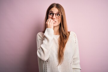 Young beautiful redhead woman wearing casual sweater and glasses over pink background looking stressed and nervous with hands on mouth biting nails. Anxiety problem.