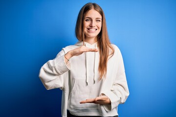 Young beautiful redhead sporty woman wearing sweatshirt over isolated blue background gesturing with hands showing big and large size sign, measure symbol. Smiling looking at the camera. Measuring