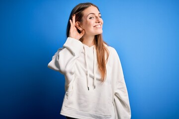 Young beautiful redhead sporty woman wearing sweatshirt over isolated blue background smiling with hand over ear listening an hearing to rumor or gossip. Deafness concept.