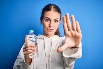 Young beautiful redhead woman doing sport drinking bottle of water over blue background with open hand doing stop sign with serious and confident expression, defense gesture