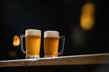 two cold mugs with beer, with overflowing foam, on wooden table and dark background, space for writing