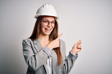 Young beautiful redhead architect woman wearing security helmet over white background smiling and looking at the camera pointing with two hands and fingers to the side.
