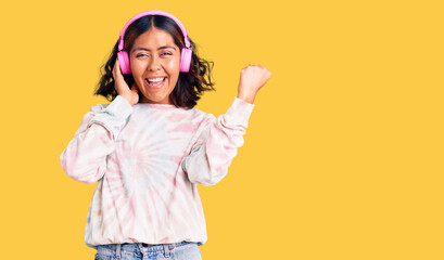 Young beautiful mixed race woman listening to music using headphones screaming proud, celebrating victory and success very excited with raised arms