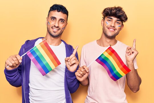 Young gay couple holding rainbow lgbtq flags smiling happy pointing with hand and finger to the side