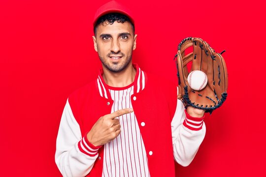 Young hispanic man wearing baseball uniform holding golve and ball smiling happy pointing with hand and finger