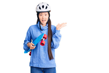 Young beautiful chinese woman wearing security helmet holding skate celebrating victory with happy smile and winner expression with raised hands