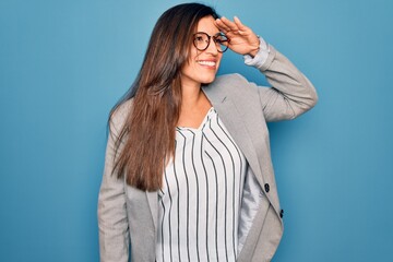 Young hispanic business woman wearing glasses standing over blue isolated background very happy and smiling looking far away with hand over head. Searching concept.
