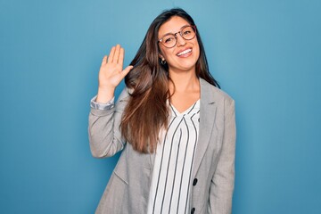 Young hispanic business woman wearing glasses standing over blue isolated background Waiving saying...
