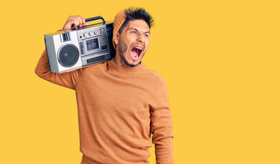 Handsome latin american young man holding boombox, listening to music angry and mad screaming frustrated and furious, shouting with anger. rage and aggressive concept.