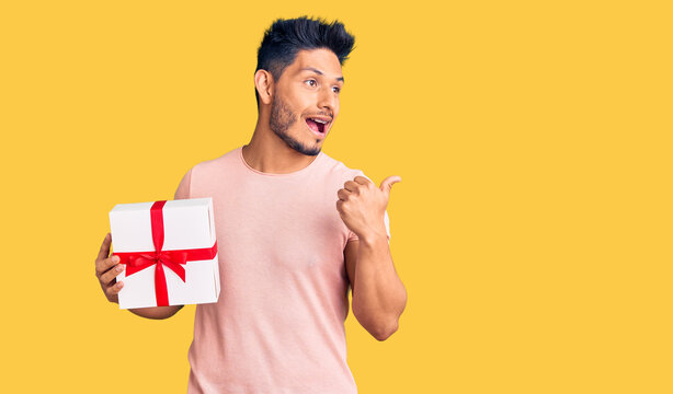 Handsome latin american young man holding gift pointing thumb up to the side smiling happy with open mouth