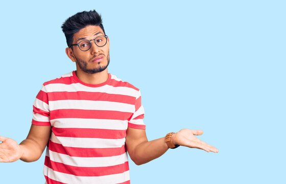 Handsome latin american young man wearing casual clothes and glasses clueless and confused with open arms, no idea concept.