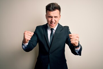 Young handsome business man wearing elegant suit and tie over isolated background angry and mad raising fists frustrated and furious while shouting with anger. Rage and aggressive concept.