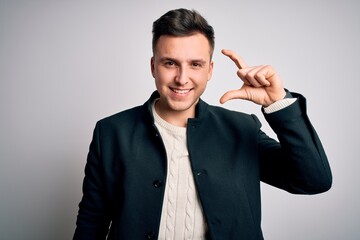 Young handsome business mas wearing elegant winter coat standing over isolated background smiling and confident gesturing with hand doing small size sign with fingers looking and the camera. Measure.