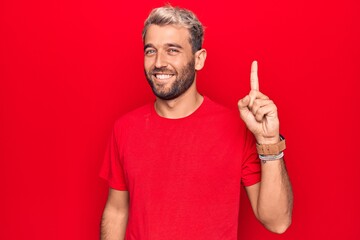 Fototapeta premium Young handsome blond man wearing casual red t-shirt standing over isolated red background smiling with an idea or question pointing finger up with happy face, number one