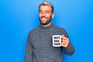 Handsome blond man drinking cup of coffee with best dad ever message over blue background looking...