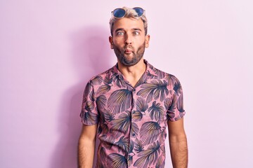 Handsome blond man on vacation wearing casual shirt and sunglasses over pink background making fish face with lips, crazy and comical gesture. Funny expression.