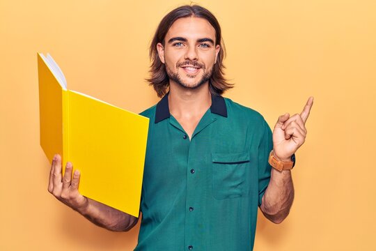 Young handsome man holding book smiling happy pointing with hand and finger to the side