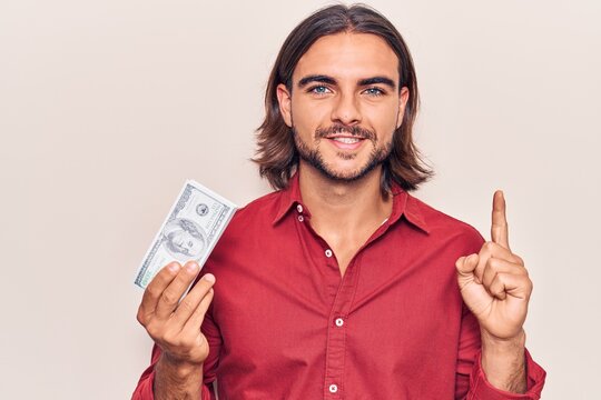 Young handsome man holding dollars smiling with an idea or question pointing finger with happy face, number one