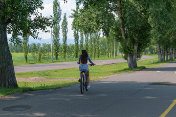 Girl is riding a bicycle. View from the back. Shorts, long hair. Asphalt road in park. Green grass and trees in summer. Concept of a healthy lifestyle, outdoor activities.